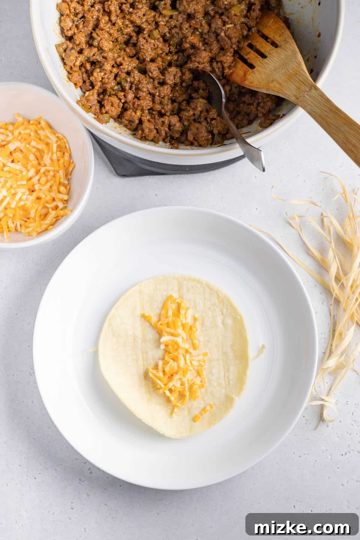 Crispy Air Fryer Beef Taquitos 6 Shredded Mexican cheese spread in a vertical line on a soft corn tortilla, placed over a corn husk strip on a white plate.
