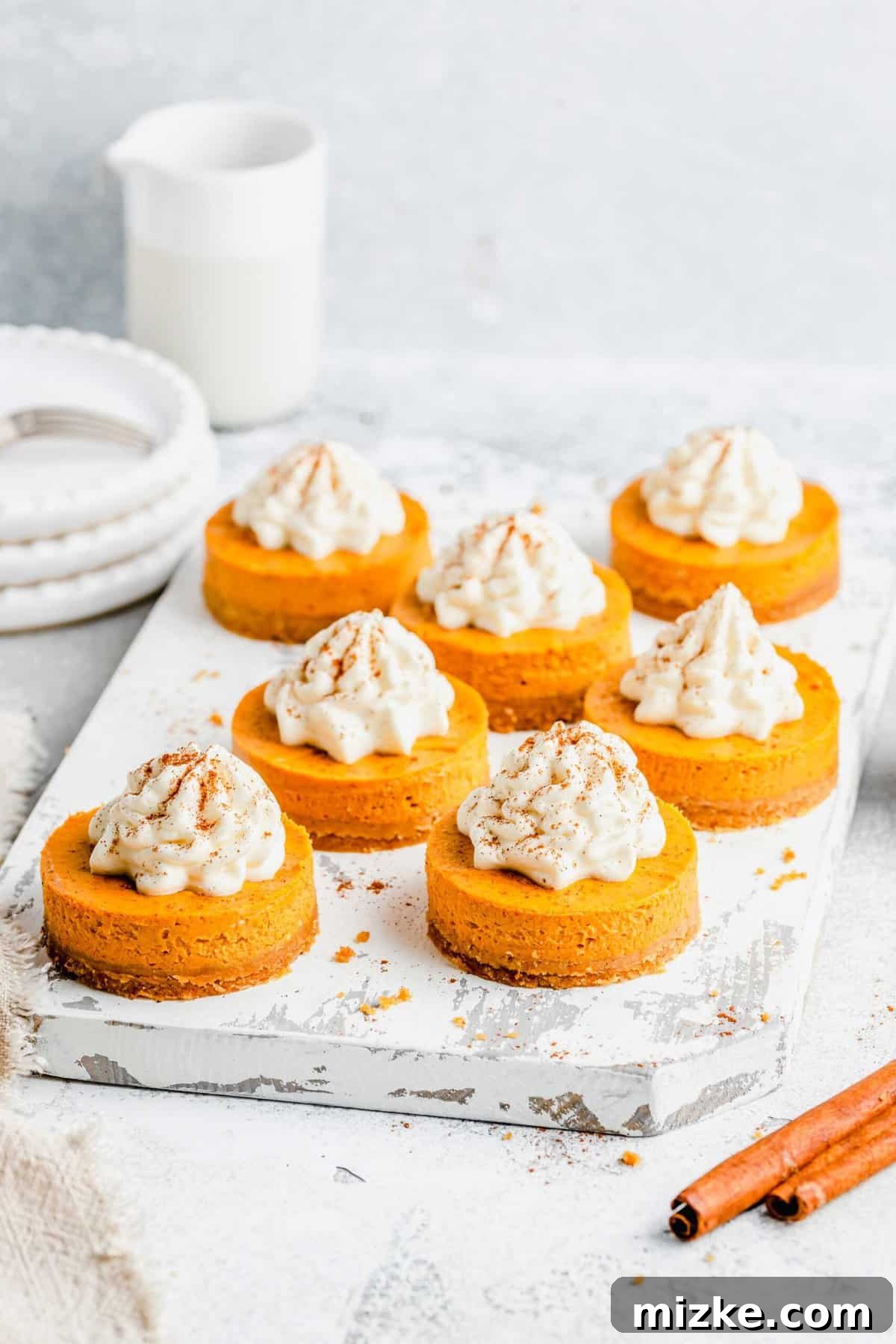Creamy mini pumpkin cheesecake bites are the perfect crowd-friendly Thanksgiving dessert filled with mouthwatering pumpkin flavor. A white tray with seven pumpkin cheesecake bites, garnished with cinnamon, with cinnamon sticks in front of the tray and plates and a pitcher behind it