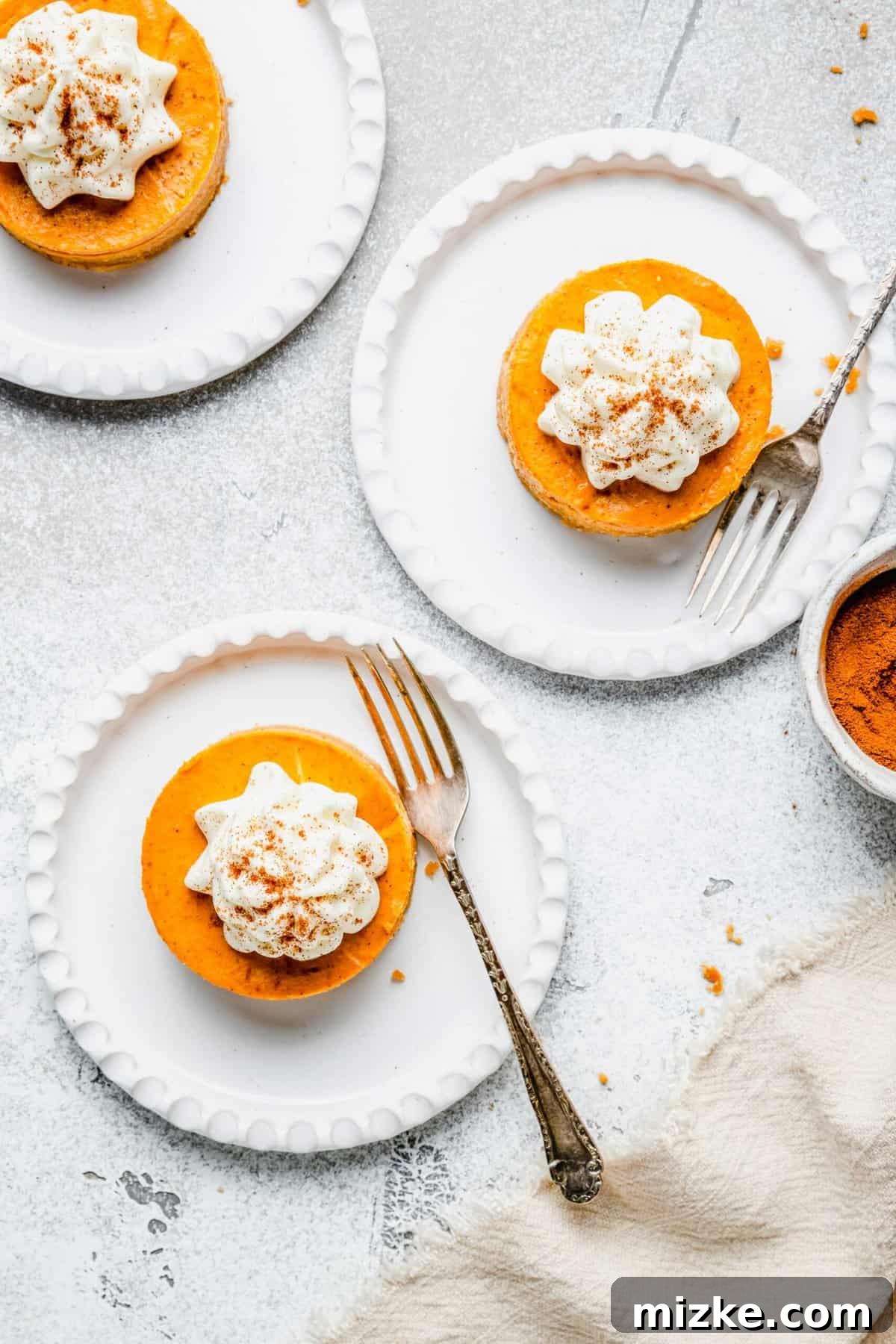 Creamy mini pumpkin cheesecake bites are the perfect crowd-friendly Thanksgiving dessert filled with mouthwatering pumpkin flavor. Three plates with a mini pumpkin cheesecake bite on them, dusted with cinnamon, with a fork on each plate, next to a bowl of ground cinnamon