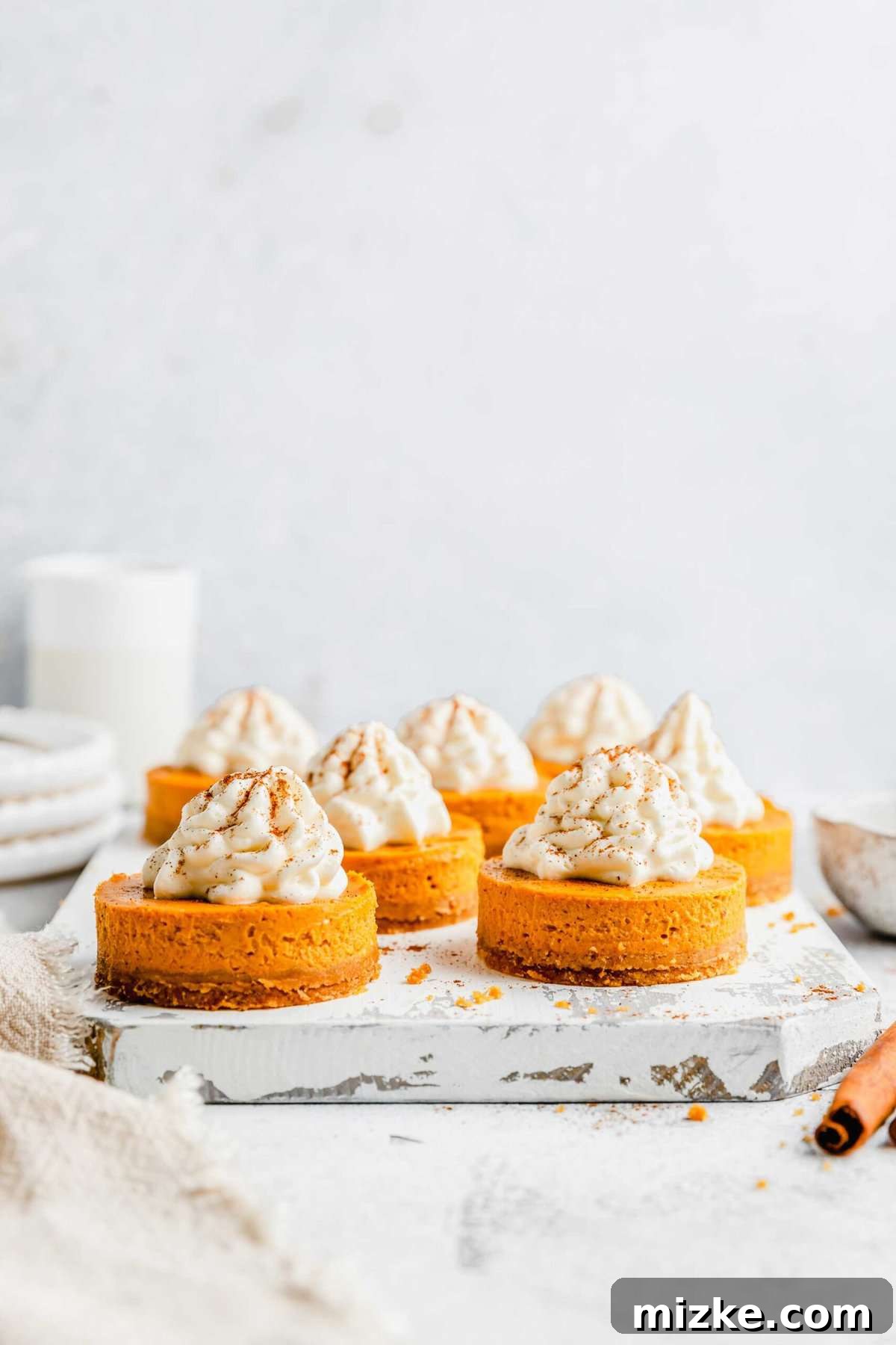 Creamy mini pumpkin cheesecake bites are the perfect crowd-friendly Thanksgiving dessert filled with mouthwatering pumpkin flavor. A tray filled with mini pumpkin cheesecake bites, dusted in cinnamon, with a tray and pitcher in the background, and a kitchen towel and cinnamon stick in the foreground