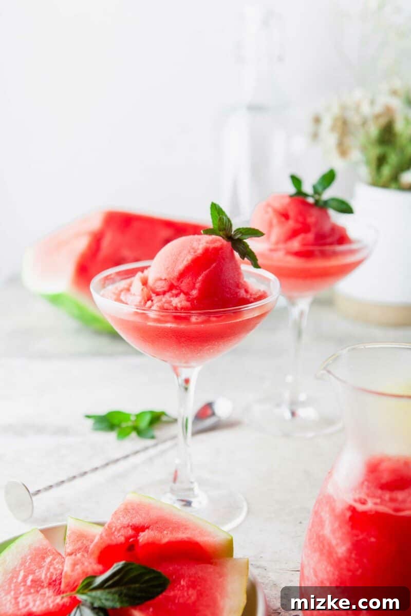 Two elegant glasses of Watermelon Frosé are presented on a table, with a large, ripe chunk of watermelon in the background, symbolizing its fresh ingredients.