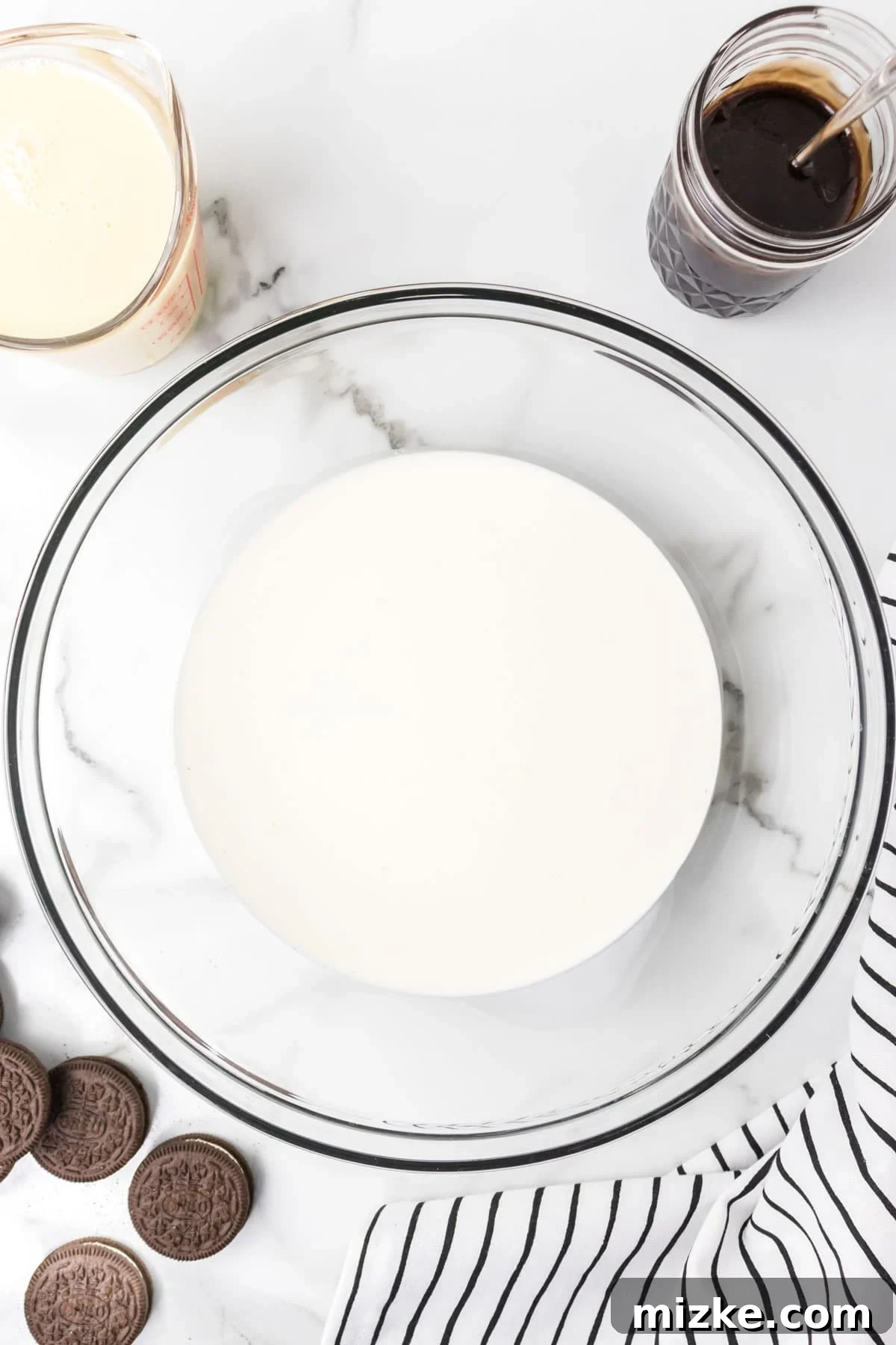 Oreo Cookie Bliss 6 Heavy whipping cream in a clear mixing bowl, next to a stack of Oreos and a striped linen towel, ready for whipping.
