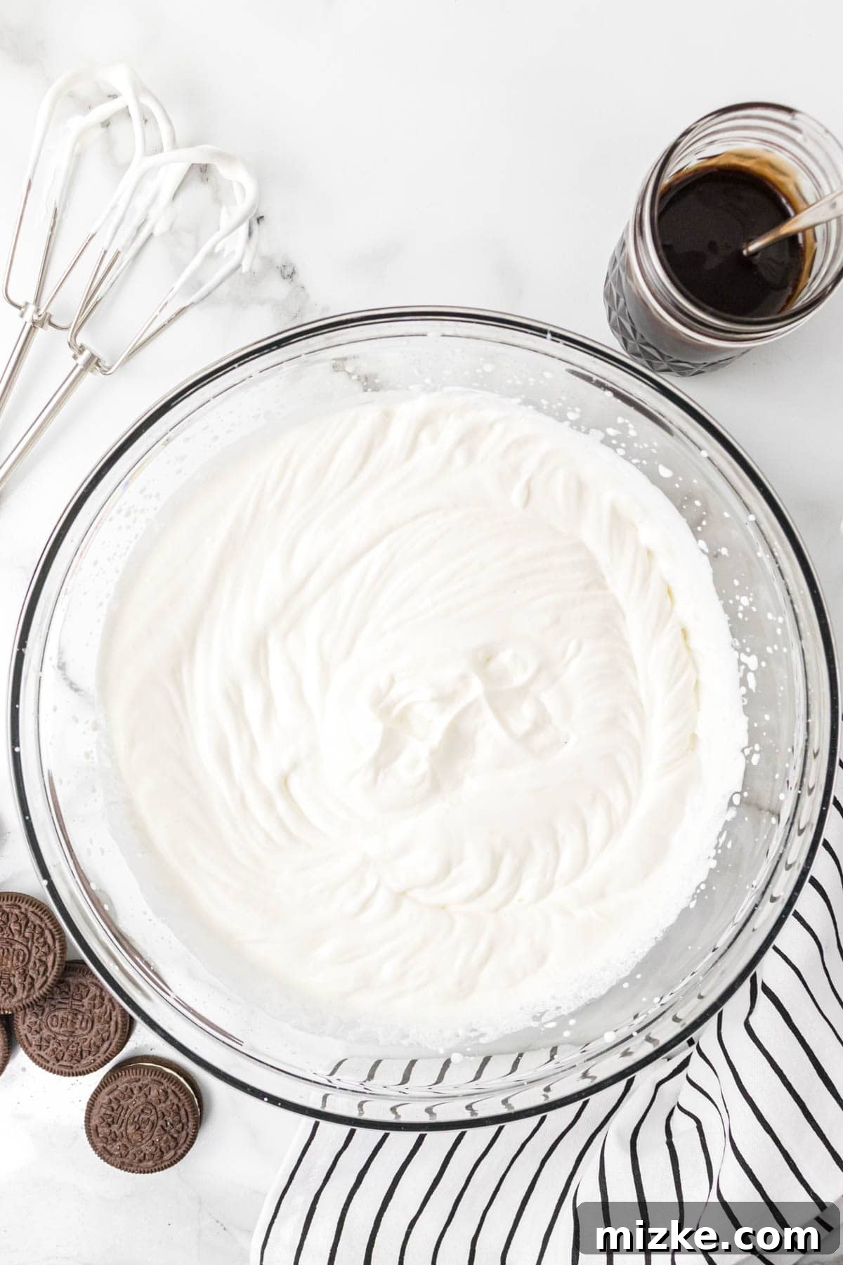 Oreo Cookie Bliss 7 Heavy whipped cream, showing stiff peaks, in a clear bowl with electric mixer beaters resting nearby.