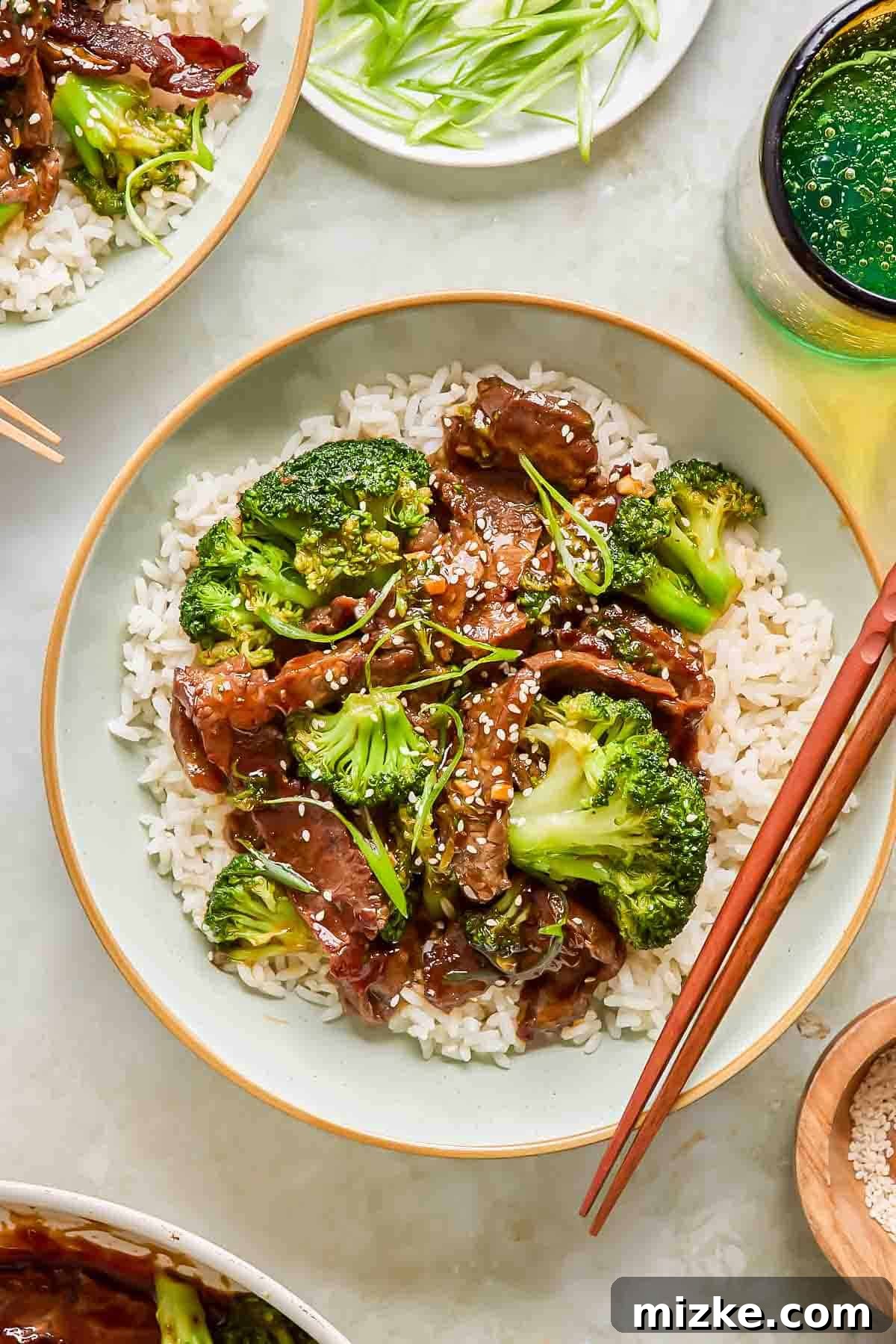 Teriyaki Beef with Crisp Broccoli 3 A beautifully plated bowl of white rice topped with generous portions of beef and broccoli, glistening with homemade teriyaki sauce, and elegantly garnished with sesame seeds and green onions, accompanied by traditional chopsticks.
