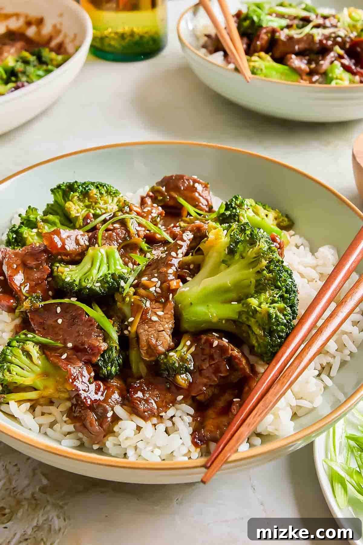 Teriyaki Beef with Crisp Broccoli 4 A rustic bowl filled with steaming white rice, topped with savory beef and bright green broccoli florets, all generously coated in a rich teriyaki sauce and sprinkled with white sesame seeds. Wooden chopsticks are neatly placed on the side of the bowl, inviting a delightful meal.