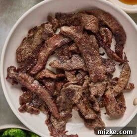 A plate of cooked beef strips sits on a table surrounded by bowls of broccoli, sauce, and honey.
