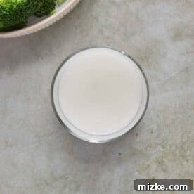 A glass of cornstarch slurry liquid next to a bowl of broccoli on a light gray countertop.