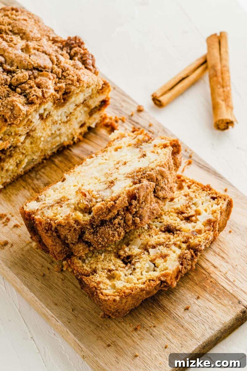 Several slices of golden-brown apple cinnamon bread, showing off their distinct swirl patterns, are arranged artfully on a light wooden cutting board.