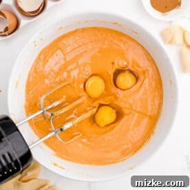 Air Fryer Pumpkin Perfection 6 A hand mixer blending orange pumpkin batter in a bowl, with three eggs alongside.