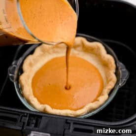 Air Fryer Pumpkin Perfection 7 Pumpkin pie filling being carefully poured from a measuring cup into a pie crust already in the air fryer.