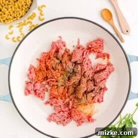 Ground beef and seasonings in a cast iron skillet next to fresh parsley and dried pasta, ready for cooking.