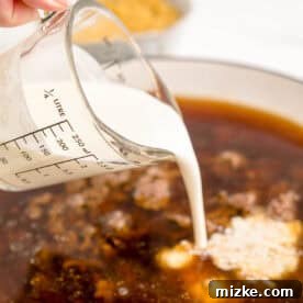 Heavy cream is being poured into a cast iron skillet with other liquids and cooked ground beef.