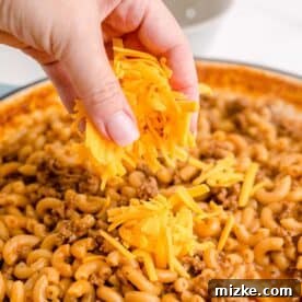 Freshly grated cheddar cheese is being sprinkled on top of the cooked beef and macaroni pasta.