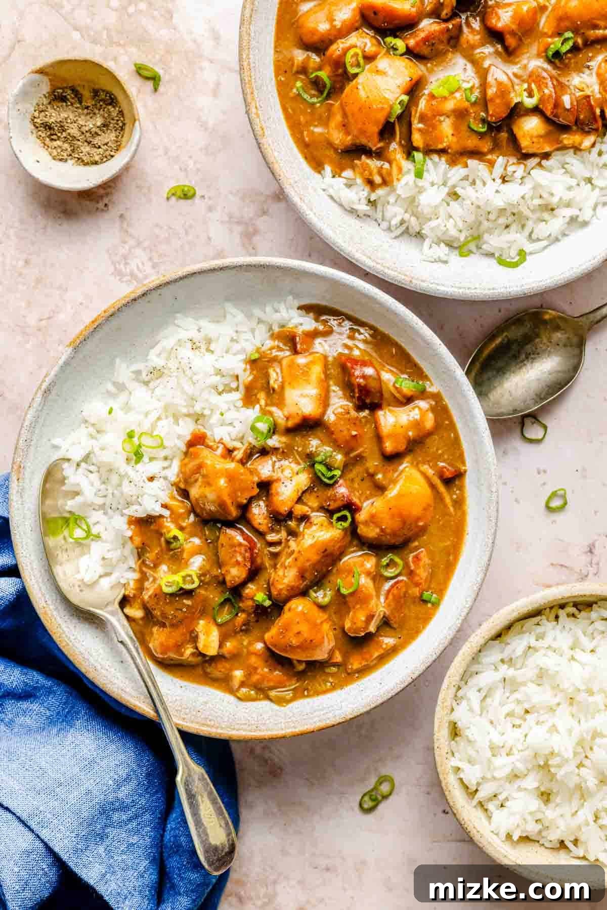 Hearty Chicken and Sausage Gumbo 2 Two bowls of richly colored chicken and sausage gumbo with white rice, garnished generously with fresh green onions. A spoon rests beside one bowl, and a small dish of black pepper is visible, adding to the inviting presentation of this classic comfort food.