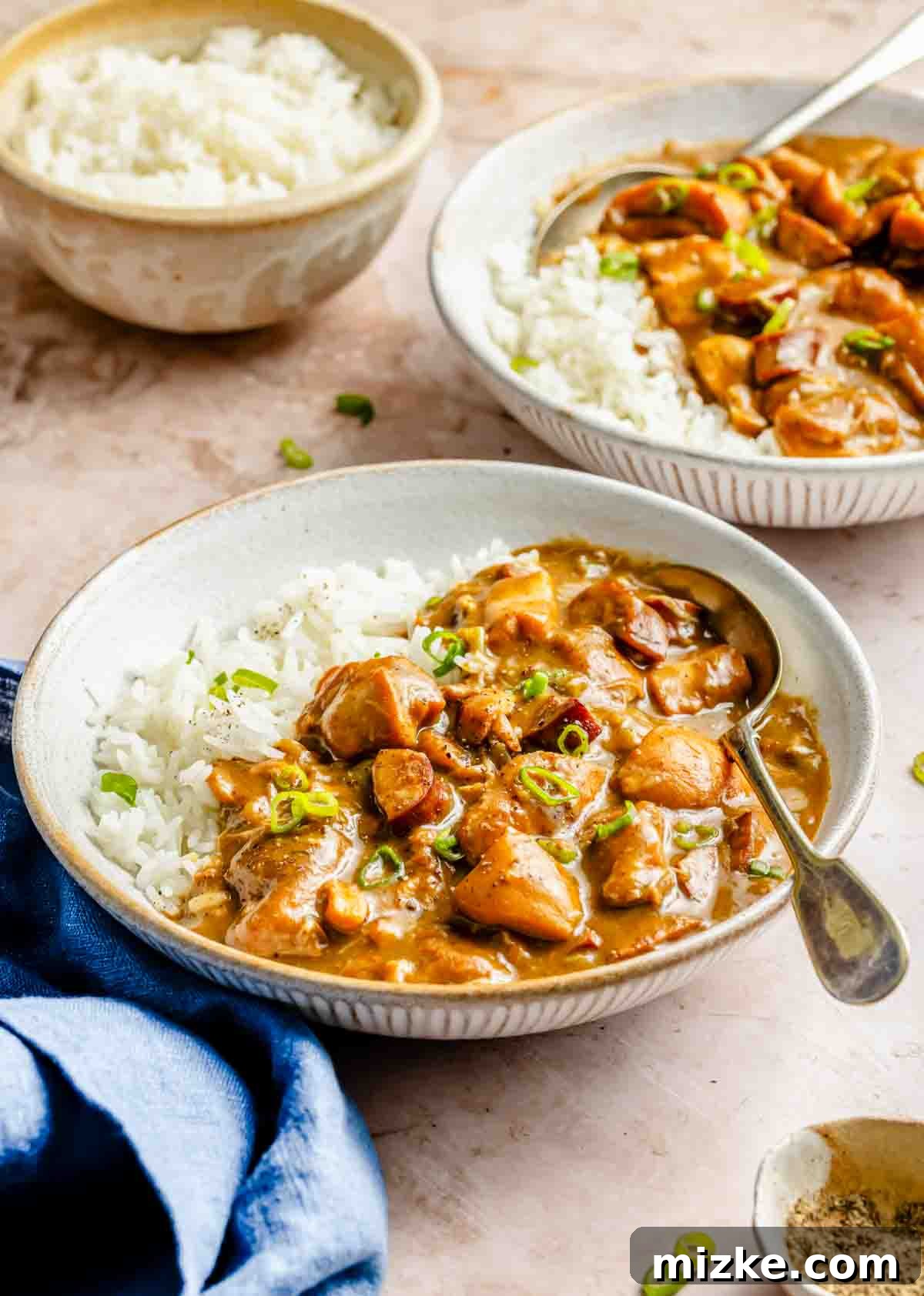 Hearty Chicken and Sausage Gumbo 3 Two inviting bowls of chicken and sausage gumbo, generously served over fluffy white rice and garnished with vibrant green onions. A small bowl of plain white rice and a neatly folded blue napkin complete the rustic table setting, evoking warmth and comfort.