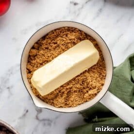 A stick of unsalted butter resting on a pile of dark brown sugar in a small saucepan before melting.