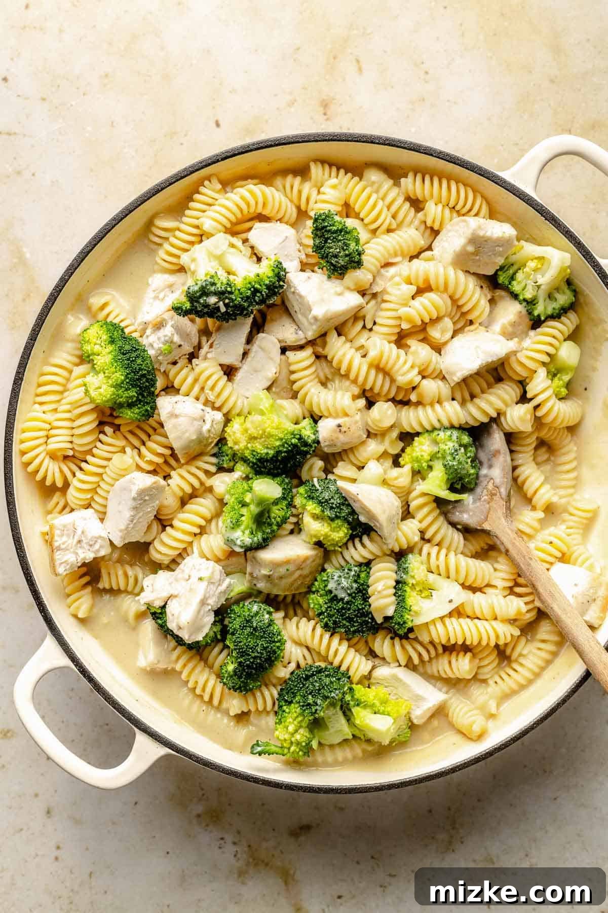 Combining cooked chicken, broccoli, and pasta with homemade Alfredo sauce in a skillet. Cooked rotini pasta, thawed broccoli, and cubed chicken being added to the creamy Alfredo sauce in a large skillet, ready to be tossed together.