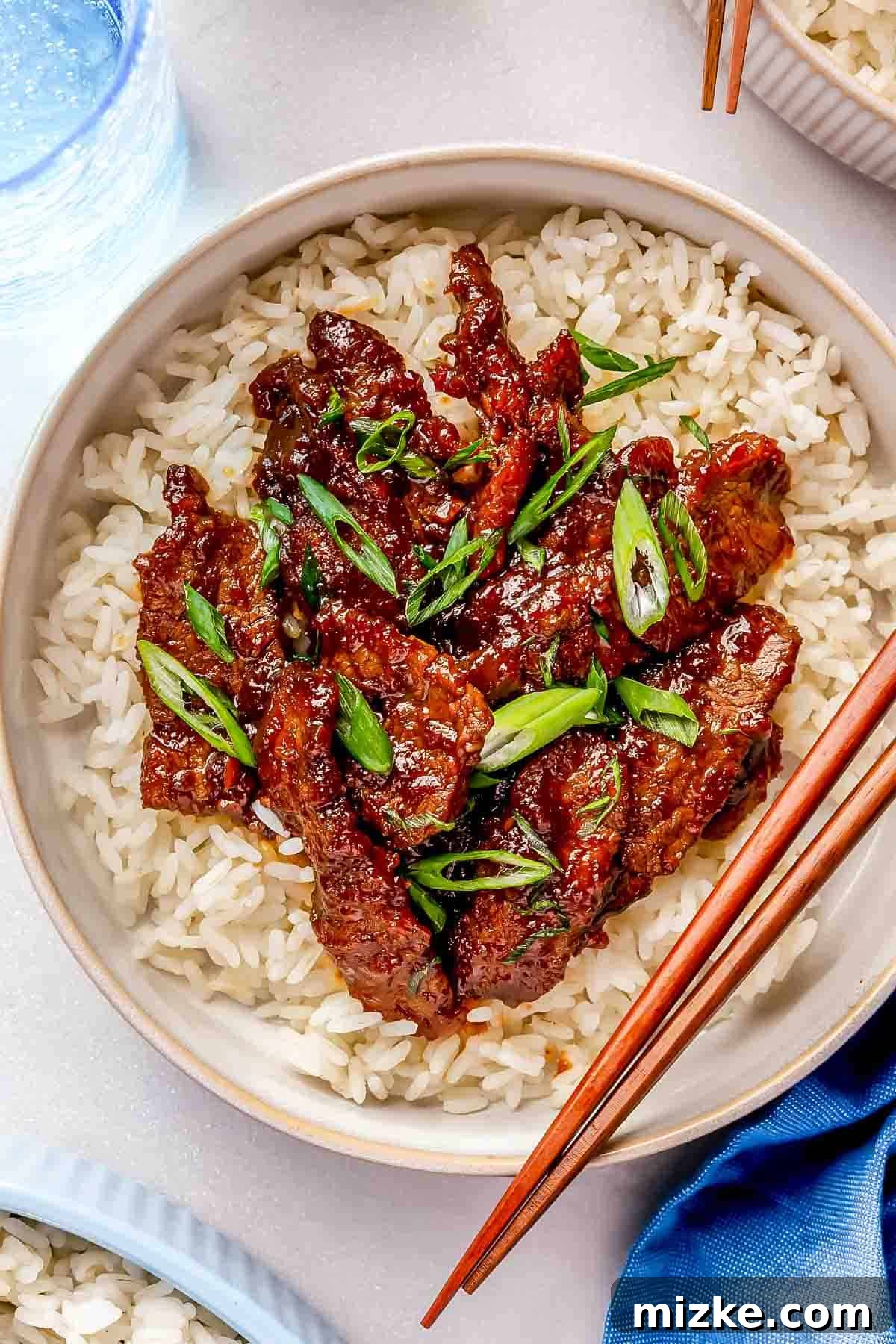 Sweet and Savory Mongolian Beef 4 A bowl of white rice topped with Mongolian beef in a dark sauce, garnished with green onions. Chopsticks rest on the bowls edge.