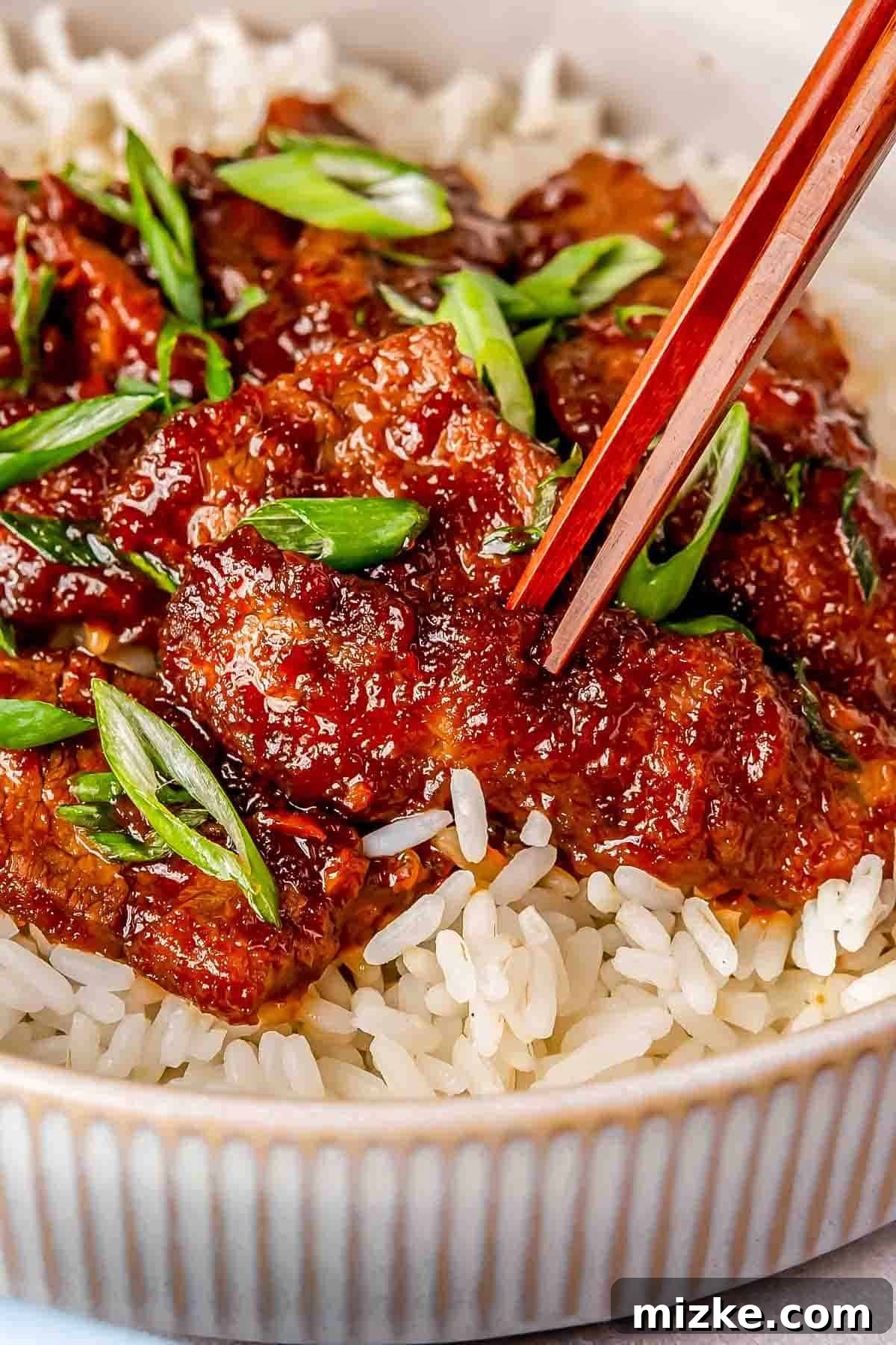 Sweet and Savory Mongolian Beef 5 A close-up of crispy Mongolian beef garnished with green onions, served over white rice in a bowl. Chopsticks are picking up a piece of beef.