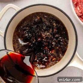 Sweet and Savory Mongolian Beef 8 Soy sauce is being poured from a measuring cup into a white pot on a countertop, surrounded by chopped green onions and a bowl of raw meat.