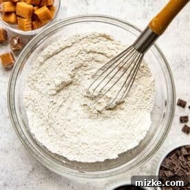 flour mixture in a clear bowl with a metal whisk next to soft caramel candies and chocolate chunks