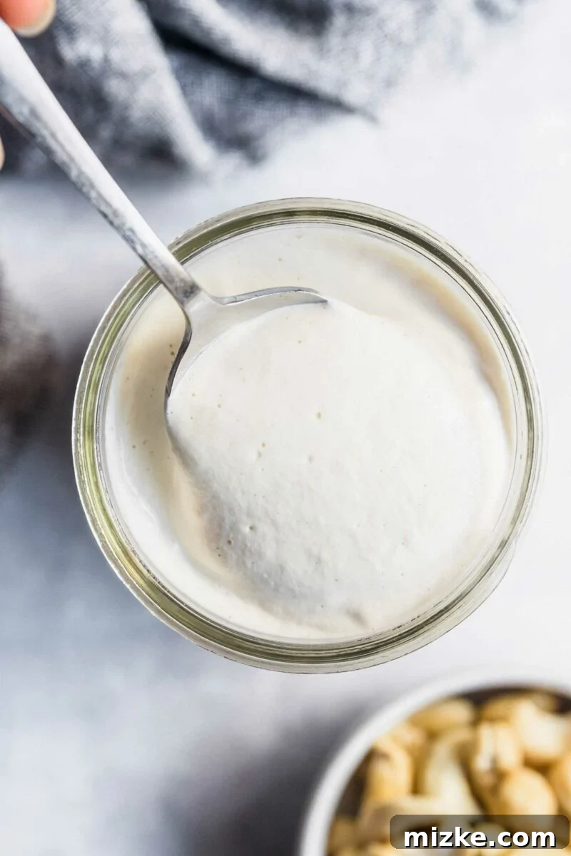Dreamy Vegan Cashew Cream 5 A silver spoon scoops a generous portion of creamy vegan cashew cream sauce from a clear glass jar.