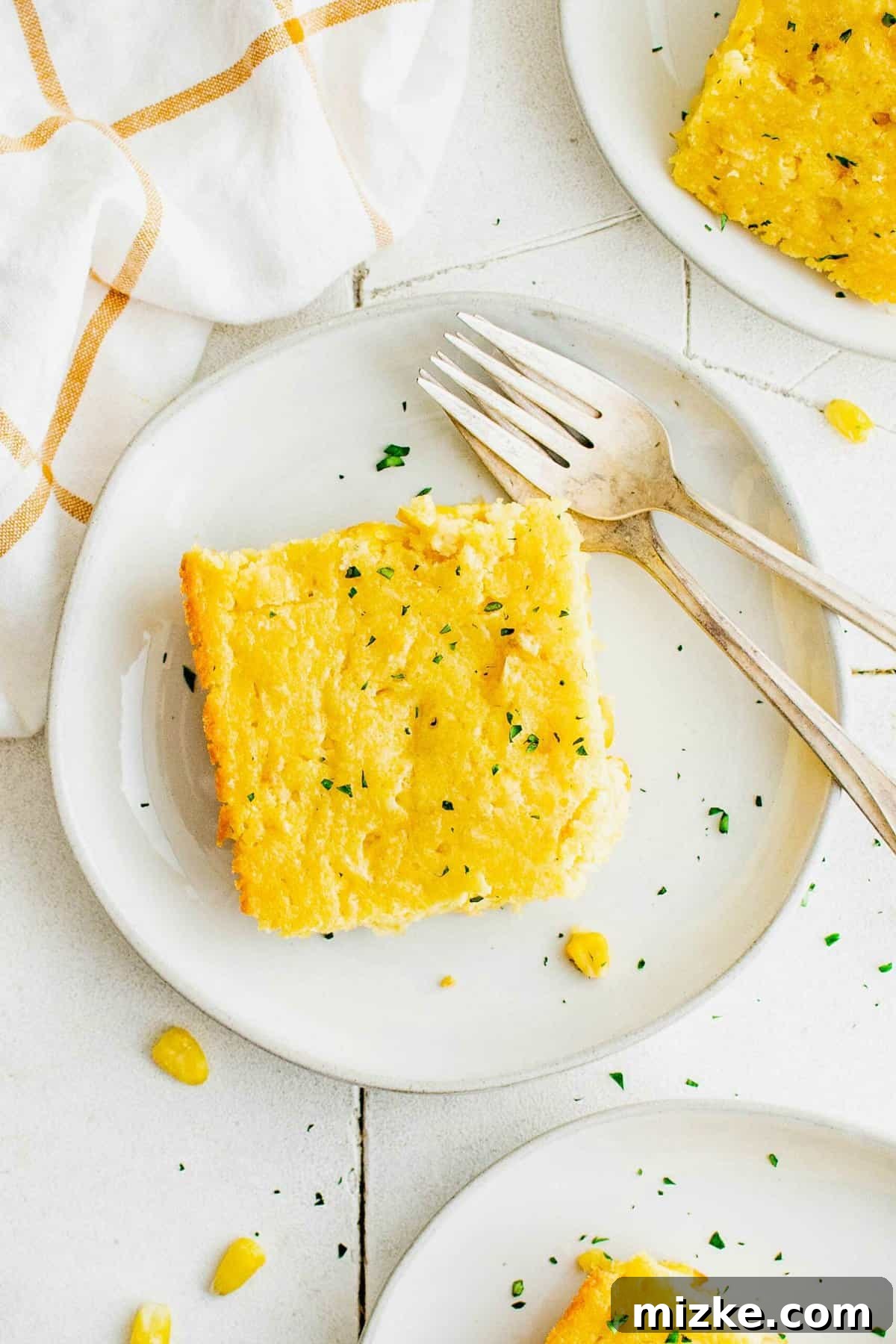 This perfectly baked cornbread casserole is a versatile and easy side dish for any meal. Two plates each holding a warm, golden slice of corn casserole, ready to be enjoyed with forks.