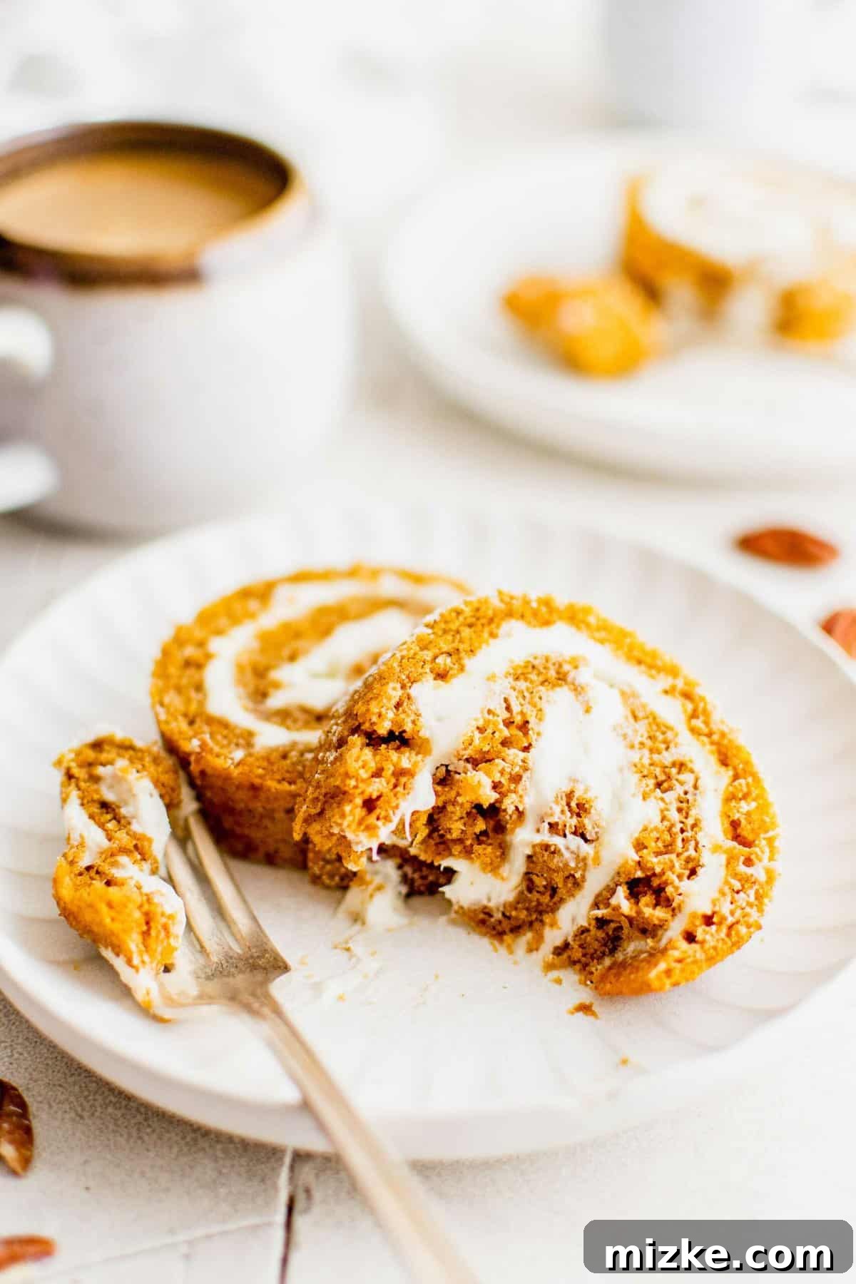 Enjoying a slice of homemade pumpkin roll with coffee. Two generous slices of pumpkin roll on a white plate, accompanied by a fork and a steaming cup of coffee in the background.