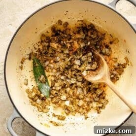 Onions, garlic, seasonings, and bay leaf in a Dutch oven with a wooden spoon.