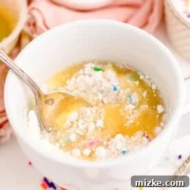 Mixing funfetti cake mix, water, and applesauce together in a white mug with a metal spoon until smooth