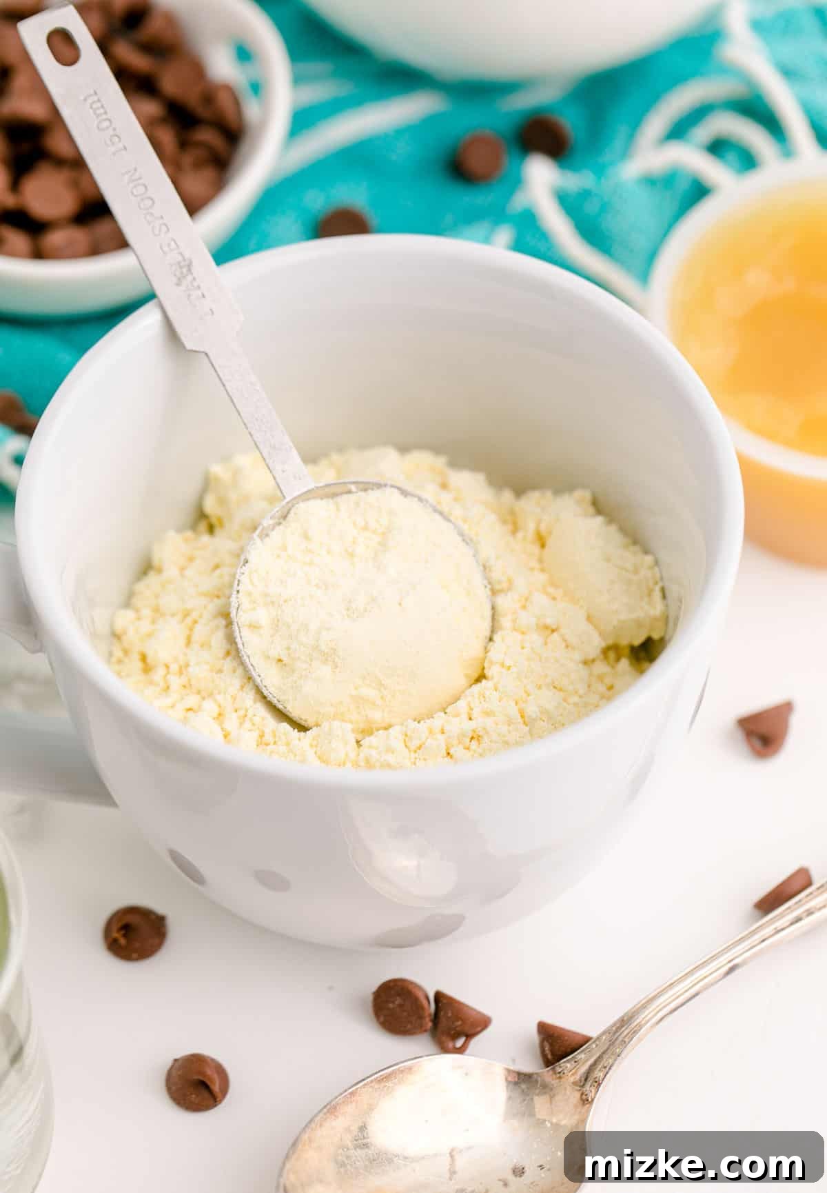 Instant Chocolate Chip Mug Treat 5 Yellow cake mix being added to a white mug with a tablespoon measuring spoon