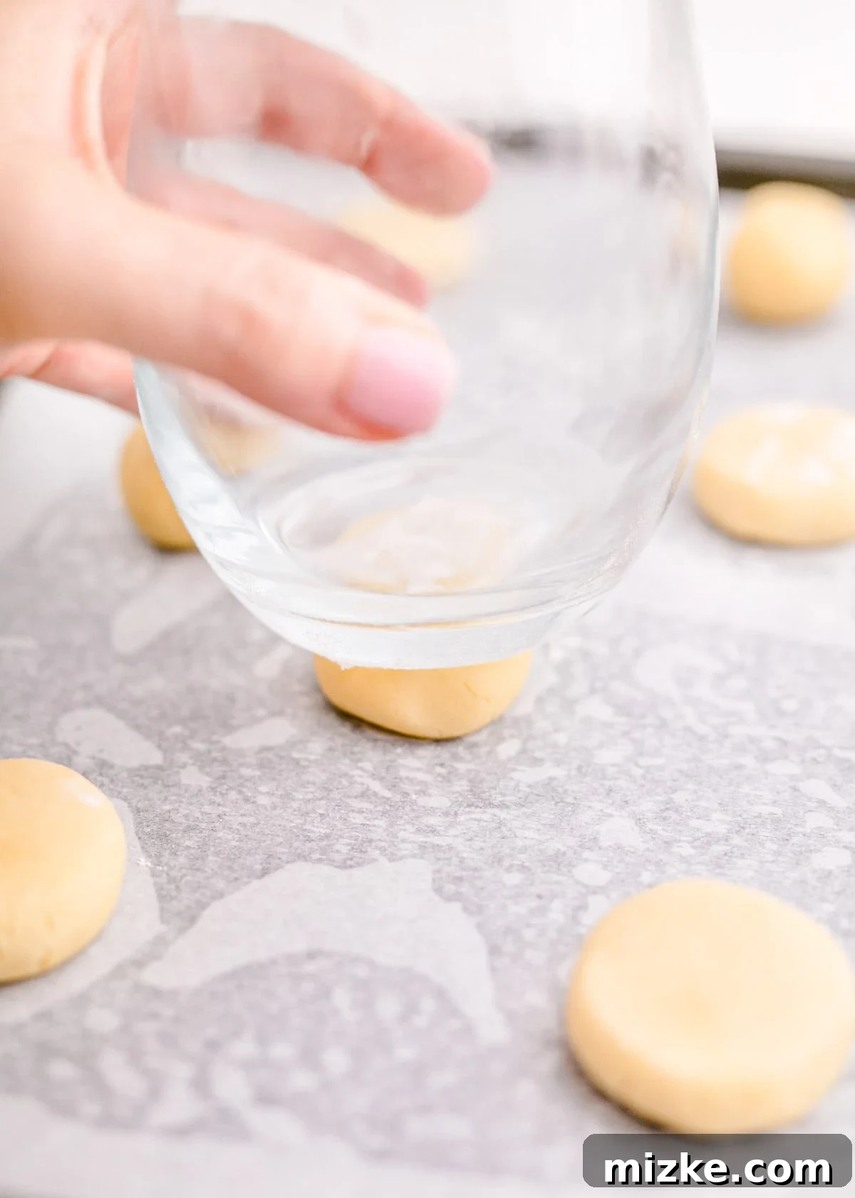 A glass is being used to gently flatten a ball of cookie dough on a baking sheet.