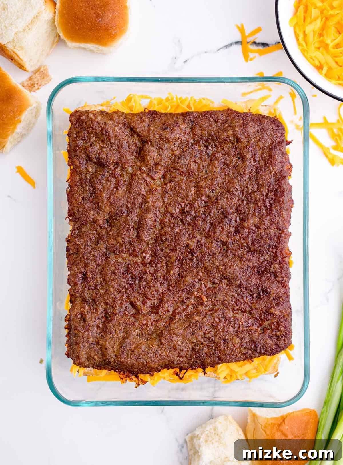 Morning Bites 12 A cooked block of pork sausage neatly placed on top of the shredded cheddar cheese layer in a glass baking dish