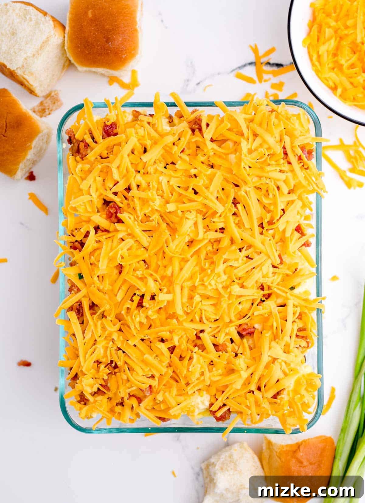 Morning Bites 15 Additional shredded cheddar cheese sprinkled over the crumbled bacon layer in a glass baking dish, preparing for the top bun