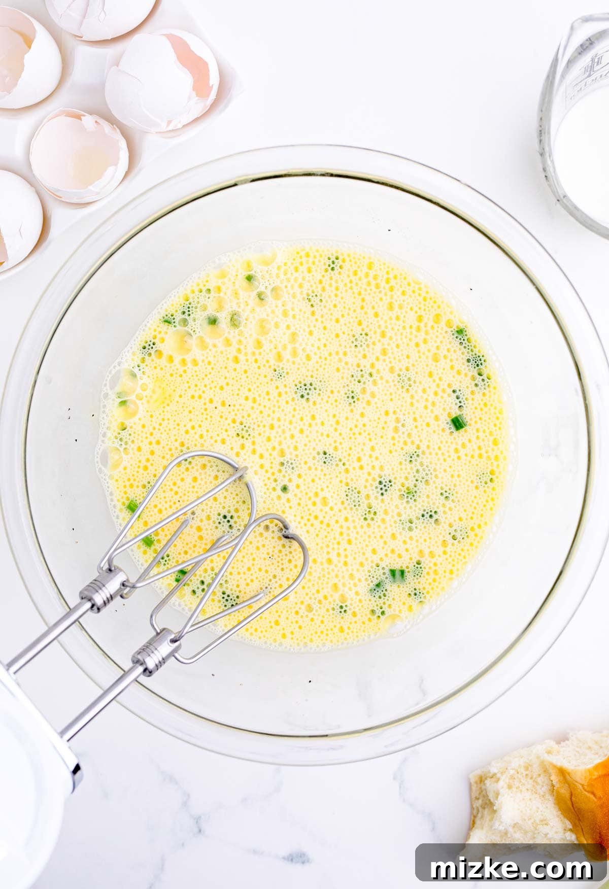 Morning Bites 9 Egg mixture in a clear glass bowl, enhanced with vibrant green onions, with an electric mixer beater inside