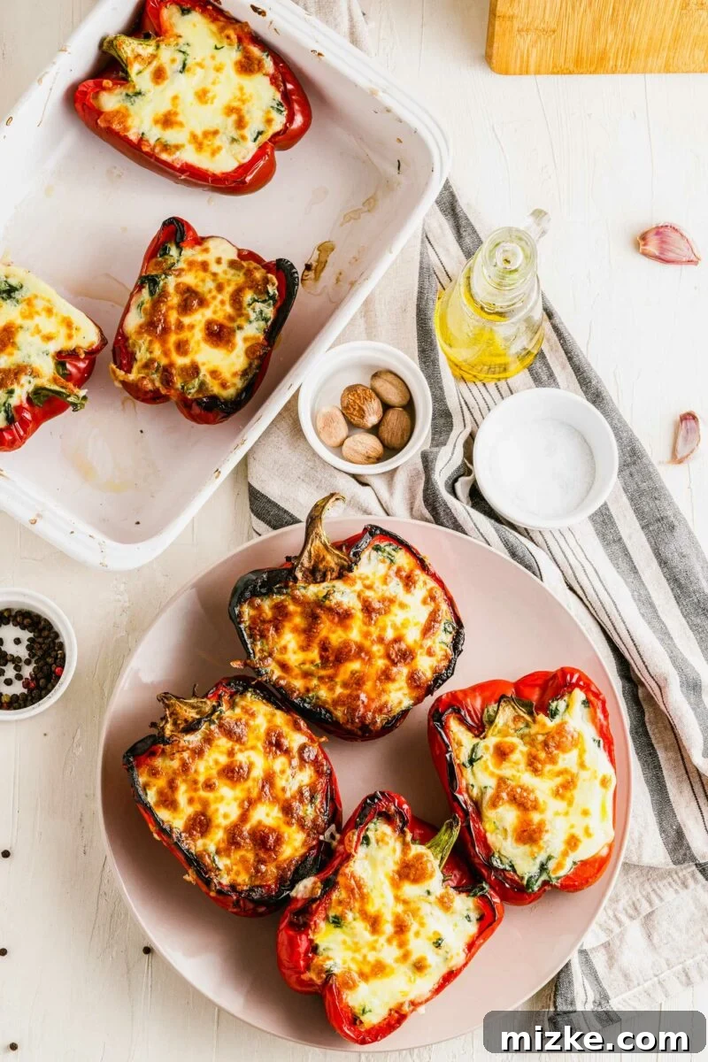 Four cooked red bell peppers, beautifully stuffed with a cheesy spinach and ricotta filling, arranged on a round white plate. A serving dish with more peppers sits nearby, inviting you to indulge.