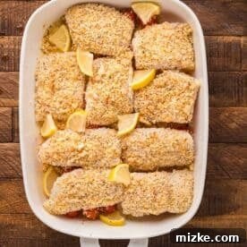 Everything bagel crusted cod baking in a dish with cherry tomatoes and fresh lemon wedges, nearly ready.