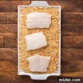 Cod fillets being coated in the panko breadcrumb mixture in a shallow dish, preparing for baking.