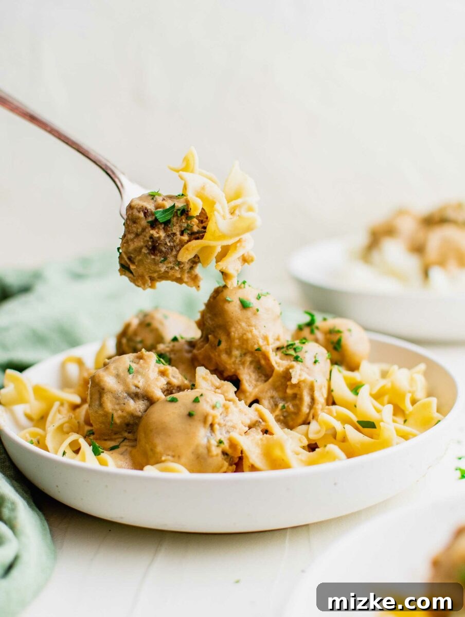 Irresistible Swedish Meatballs 5 A single, perfectly cooked meatball, tender and juicy, is being lifted by a fork along with a few strands of egg noodles from a bowl, highlighting the enticing texture and sauce.