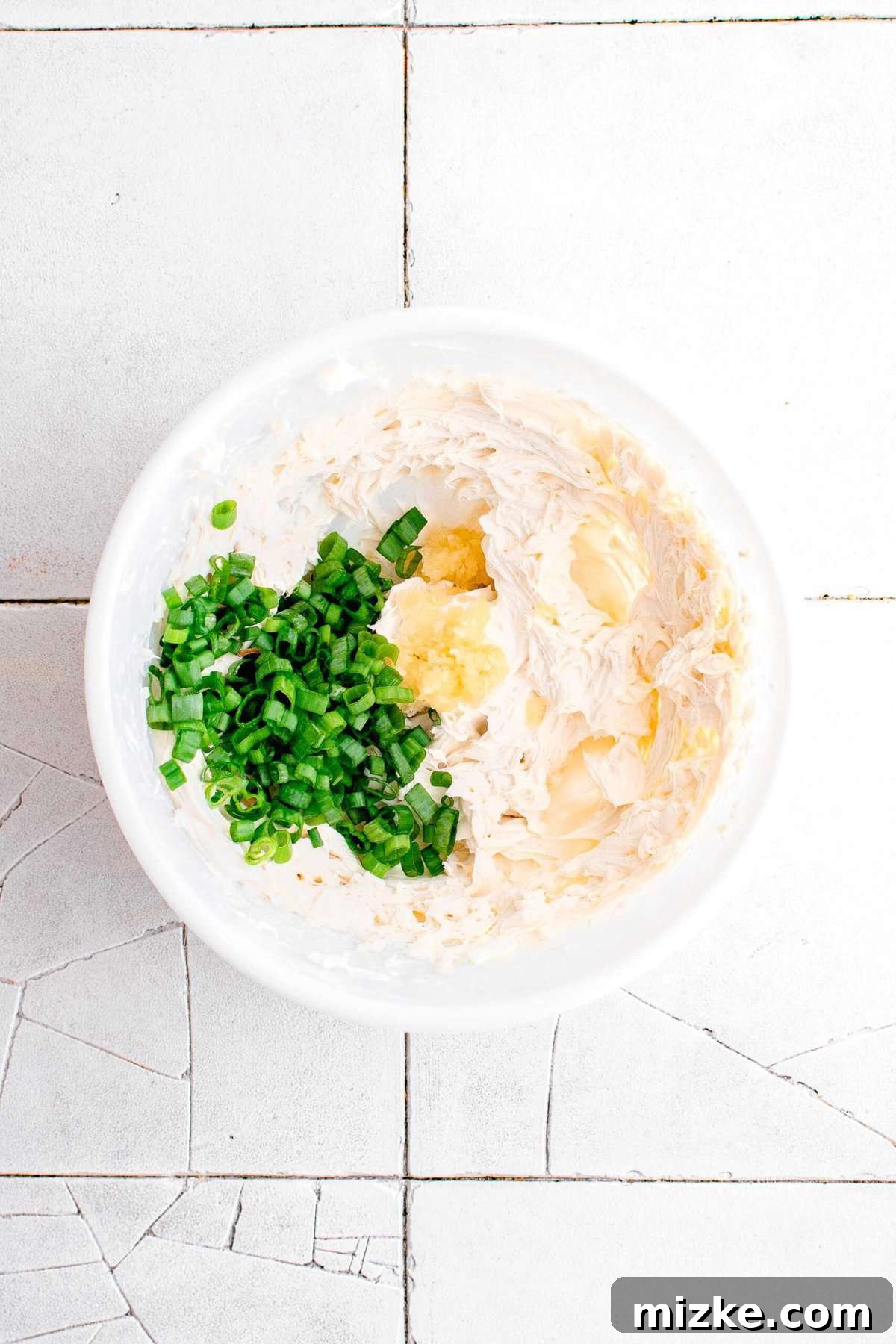 Golden Crispy Crab Bites 6 A creamy, whipped cream cheese mixture in a white bowl, with chopped scallions and minced garlic being added, set against cracked grey tiles.