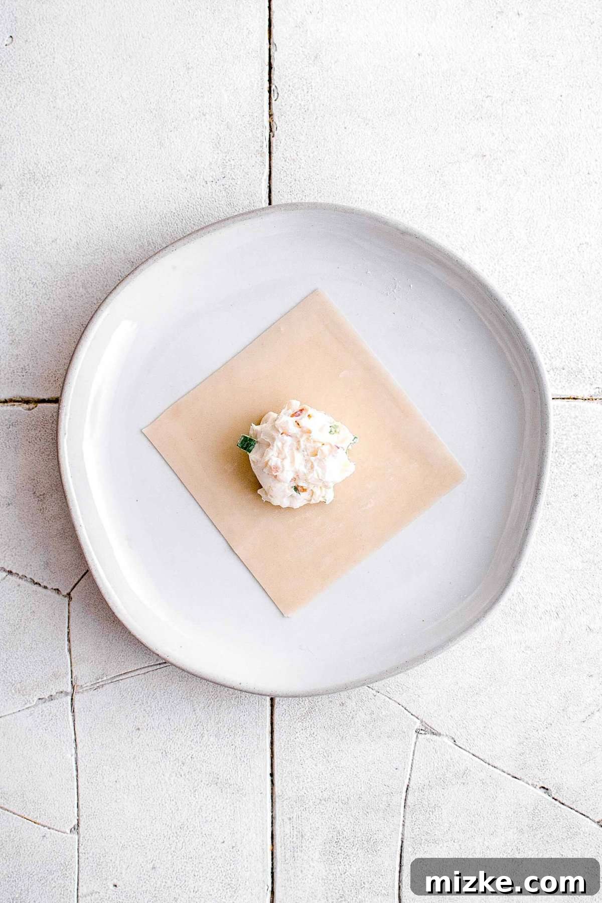 Golden Crispy Crab Bites 8 Two teaspoons of creamy crab rangoon filling meticulously placed in the center of a square wonton wrapper on a white plate, resting on cracked grey tiles.