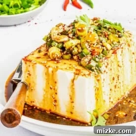 A block of tofu is topped with green onions, cilantro, sesame seeds, and a homemade oil.