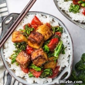 A serving of spicy tofu stir fry sits on a bed of white rice in a small bowl. Chopsticks sit on the side of the bowl.