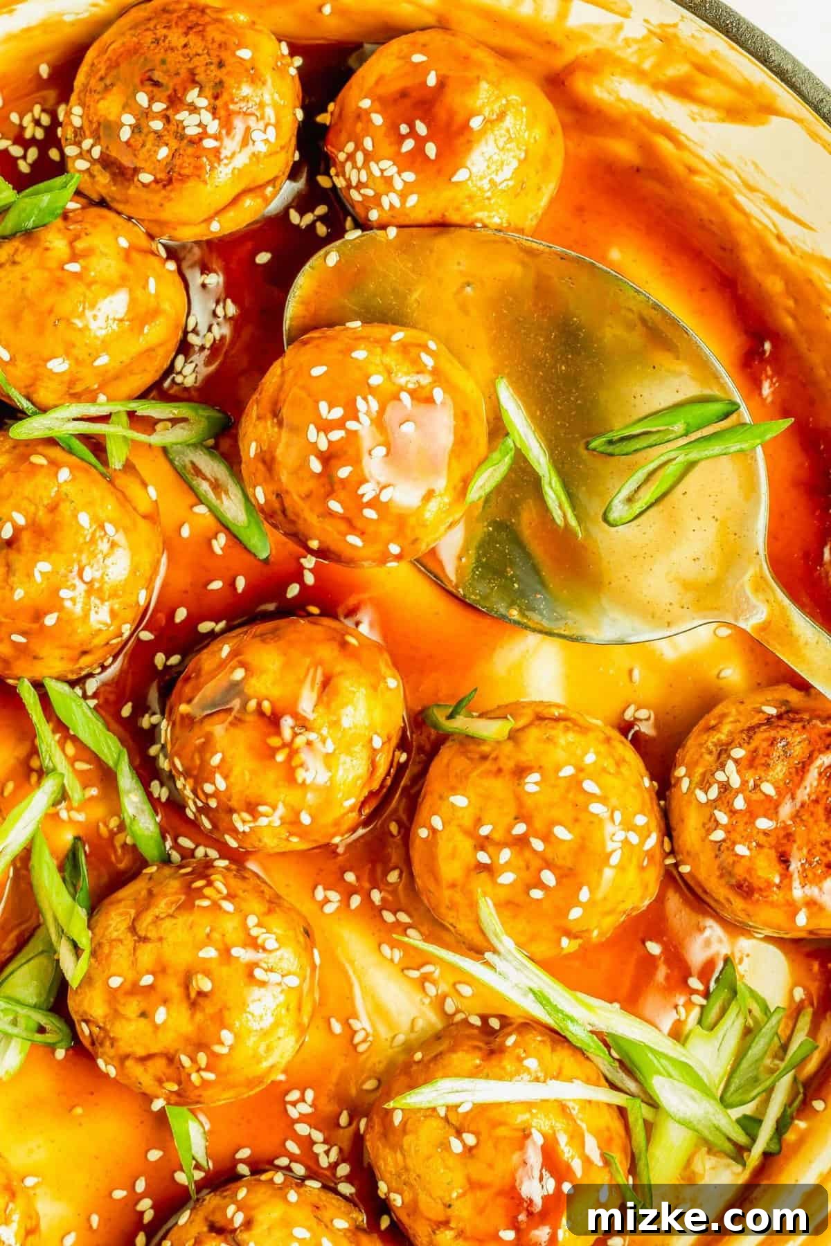 Perfectly glazed teriyaki tofu meatballs, ready to serve. Overhead view of teriyaki-coated tofu meatballs in a black enamel skillet with a large serving spoon, garnished with scallions and sesame seeds.