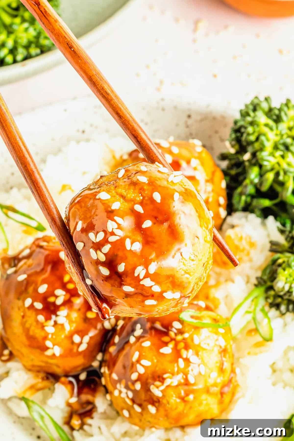 A perfect bite of teriyaki tofu meatball. Chopsticks picking up a glistening teriyaki tofu meatball adorned with sesame seeds, against a blurred background.