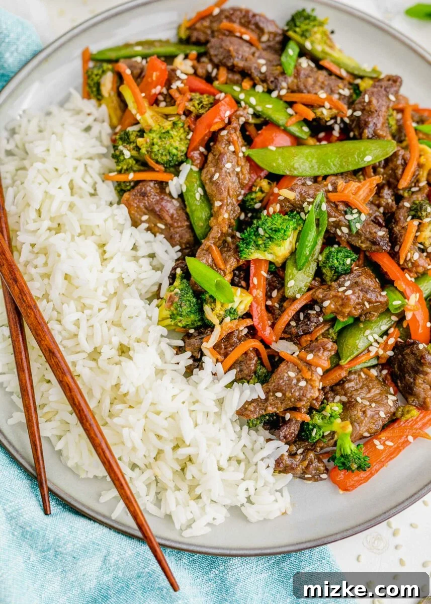 Sizzling Beef Stir Fry 5 A beautifully presented bowl of white rice topped with generous portions of beef stir fry, garnished with fresh green onions and sesame seeds, with chopsticks poised for eating.