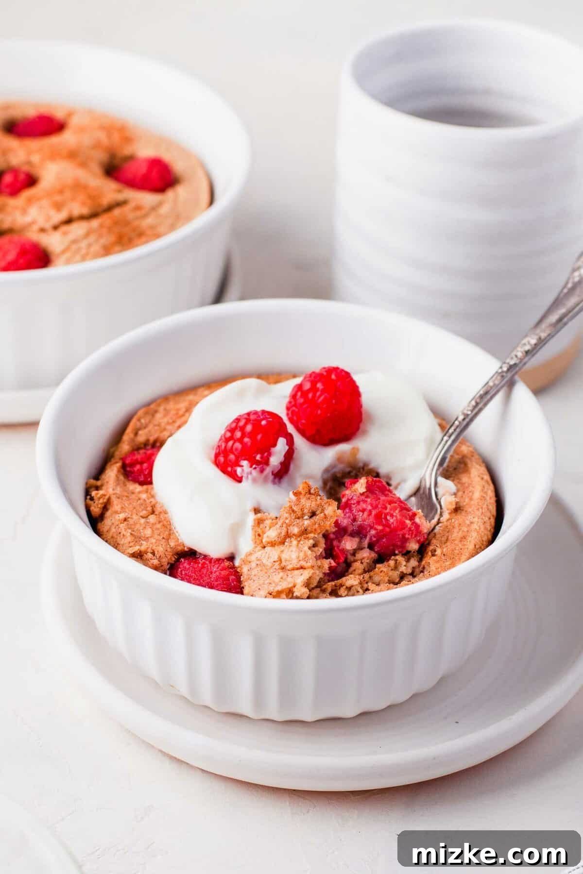 Raspberry Oatmeal Bake 2 Raspberry baked oatmeal topped with creamy Greek yogurt and vibrant fresh raspberries, served in a pristine white bowl with a spoon, inviting you to indulge in a wholesome breakfast.