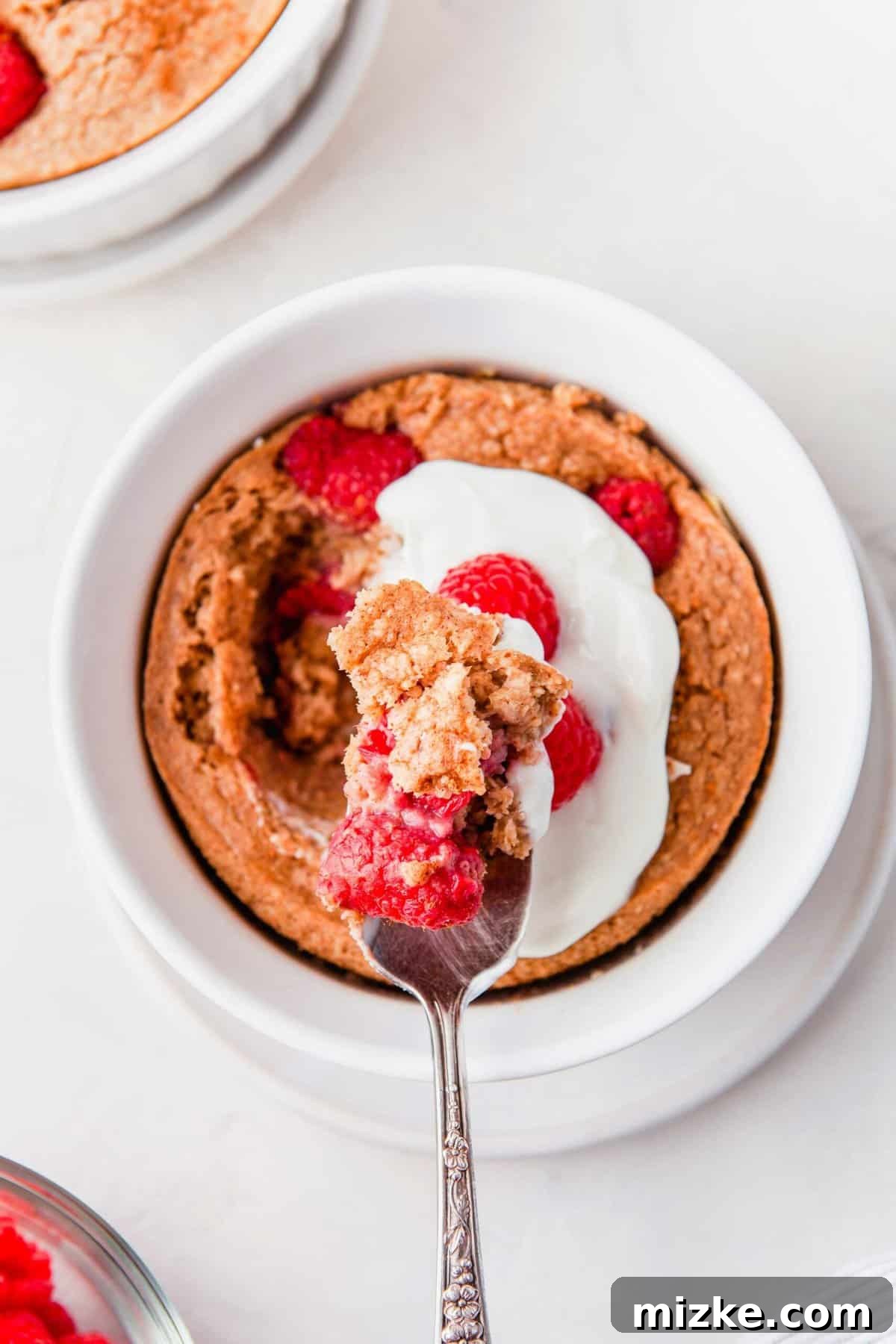 Raspberry Oatmeal Bake 4 An overhead shot of raspberry baked oatmeal in a white bowl with a spoon, perfectly baked and adorned with fresh berries, showcasing its ideal cake-like texture.