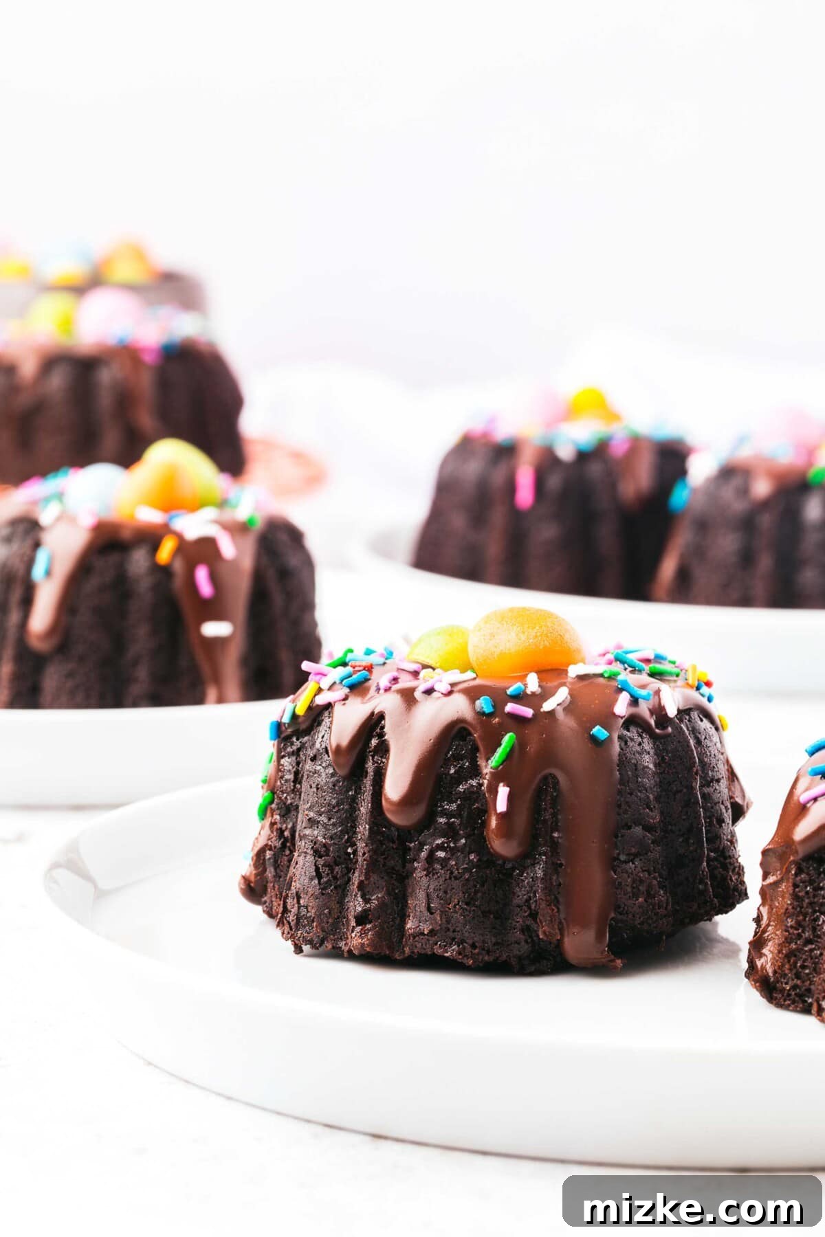 A single mini bundt cake, elegantly topped with glossy ganache, festive sprinkles, and two charming chocolate eggs, presented on a minimalist white plate.