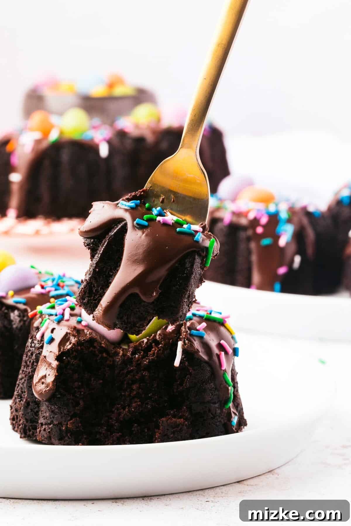 A single bite of a mini bundt cake, showing its moist interior and ganache topping, is being lifted from a plate by a fork.